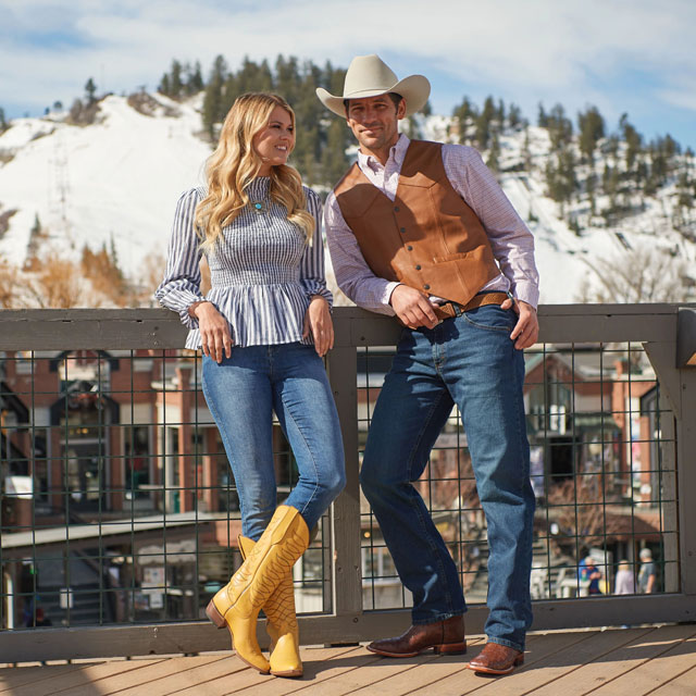 A man and woman standing on a patio wearing cowboy boots. The woman is wearing a blouse shirt and jeans while the man is wearing a shirt, vest, jeans, and cowboy hat.